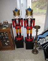 Two tall wooden nutcracker figures beside a tall brass candleholder, all standing on floor near window.
