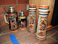 Photo showing four German beer steins arranged on a brick floor near wooden furniture. Two taller steins on right with vertical orange and blue hand-painted panels. Left side shows two shorter steins with metal lids and detailed designs.