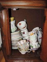 Wooden cabinet containing stacked bone china teacups and saucers with varied floral patterns and gold trims.