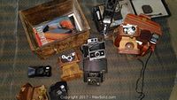 Top-down view showing collection of vintage cameras and accessories including wooden crate with manuals, Argus camera in tan leather case, black Polaroid camera, folding cameras, and light meter.