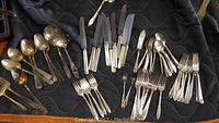 Various sterling silverware pieces displayed on a dark quilted surface including spoons, knives, forks, and serving utensils arranged loosely.
