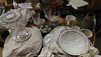 Several antique china plates and bowls with floral designs and gold trim, surrounded by drinking glasses and ceramic mugs on a table.