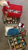 Overview of mid century glass Christmas ornaments, blue bead garland, and storage bin with Christmas hat.