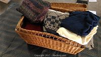 Wicker basket filled with assorted sweaters and folded clothes, placed on carpeted floor
