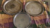 Three sterling silver platters arranged on a patterned fabric background. Two platters have ornate etched floral designs with decorative rims; one platter is plain with visible tarnishing.