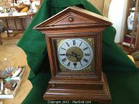 Front view of the wooden mantle clock showing triangular pediment top, clock face with Roman numerals, ornate brass detailing, and wood grain finish.