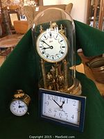 Full view of 3 clocks: mechanical anniversary clock under glass dome, small quartz alarm clock with blue frame, and small mechanical alarm clock with brass bell on top.