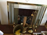 Wide angle photo showing the brass fireplace screen, fireplace tool set with stand, brass container holding a broom, and metal ash pail, placed in front of a fireplace.