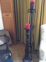 Photo showing the two floor candle stands placed side by side in front of yellow curtains and next to a beige couch, red candles placed on top of each stand.