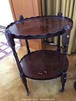 Full view of the two-tier wooden side table with scalloped edges and casters, showing the overall shape and structure.