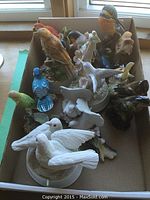Box containing sixteen various bird figurines of ceramic and bone china, mostly birds on bases with some perched on flowers and leaves.