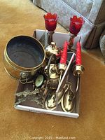 Photo showing a group of brass items including candle holders with red glass tops and a small clock in a heart-shaped brass frame, a brass flower pot, and an ornate decorative stand in a cardboard box on carpet.
