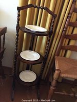 Three-tier dark wood stand with spiral turned legs and white decorative plates on each shelf, shown from front angle with window curtains in background