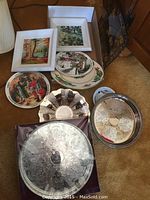 Two framed paintings by Kathleen Mudge along with several decorative plates and silverware trays shown grouped together on carpeted floor.