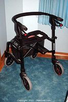Front angled view of black metal folding walker showing frame, wheels, padded seat, hand brakes, and basket underneath