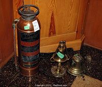 Fire extinguisher, hanging brass bell, and brass table lamp grouped on floor against wood paneling.
