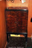 Front view of wooden bar cabinet with dark tiger stripe veneer pattern on closed double doors. Cabinet elevated on black painted legs with decorative cutouts.