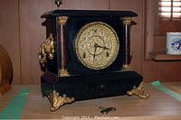 Front view of the antique Sessions mantel clock on wooden surface, showing dial and ornate gold feet, includes winding key placed in front.