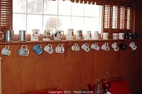 Full view of various mugs hanging and standing on a wooden shelf under windows with shutters, showing a collection of 30 beer steins and mugs in ceramic and metal.