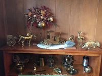 Photo showing a collection of brass figurines and decor including horses with carts, deer, fish, floral holder, mug, bell, ship, and candle holder on a wooden surface.