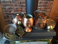Wide view of all items arranged on black surface showing kettles, pot, frypan, goblet, brass horse brasses, and decorative brass plate.