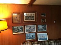 Wall display of seven framed lithograph prints and two smaller religious prints hung on wood-paneled wall above couch.