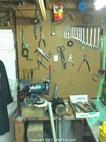 Wide view of workspace showing the bench grinder on a wooden bench, pegboard with hand tools hanging, including wrenches, pliers, scissors, and clamps