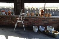 Old horse yoke, metal step ladder, wooden baskets, and assorted pots on porch floor near brick wall under window