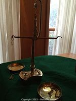 Full view of brass balance scale with two hanging pans and several weights on a green felt surface near a window.