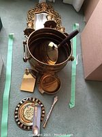Brass bucket with wooden handle, small decorative plate, two ornate brass spoons, ashtray, and belt buckle arranged on floor.