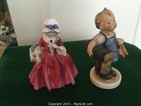 Two figurines displayed on green cloth; left is a Royal Doulton 'Lavinia' figurine in red dress, right is a Hummel figurine of a young boy holding a coat and shoes.