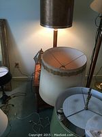 Four table lamps with shades in various decorative styles including dark brown faux leather shade and white fabric shade with floral trim, placed on a table next to a wall outlet and other furniture.