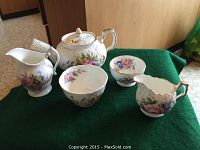 Full view of tea set on green cloth showing teapot, sugar bowl, creamer, and teacup with floral patterns and gold trimming