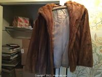 Front view of short brown fur coat with hood, partially open to show lining and label on hanger