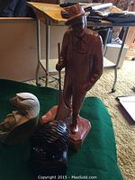 Photo showing three carved wooden statues including a standing man with rifle, a carved bird head with human face, and a smaller bird sculpture.