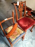 Two older wooden chairs; foreground chair has needlepoint seat with floral and musical scene, broken wooden back piece lying across seat. Background chair carved wood with red velvet cushion.