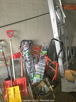 Photo showing the two aluminum ladders, red hand dolly, red shovels, broom, coiled orange extension cords, and other garden tools arranged against a wall.