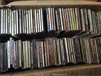 Photo showing approximately 110 assorted music CDs in jewel cases arranged vertically within a cardboard box