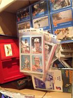 Photo of box filled with assorted sports cards including a 1989 Topps page showing four baseball players, various loose cards, and a red plastic carrying case for cards.