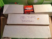 White cardboard boxes containing baseball cards with one box labeled '1981 Topps Baseball' and a retail wax box labeled 1983 Topps Baseball cards on top