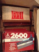 Original boxed Atari 2600 system with joystick, stacked with other vintage game boxes inside a cardboard container.
