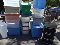 Wide view of multiple stacked plastic storage bins in various colors and two black drawer units outdoors.