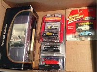 Photo shows six diecast cars in original packaging placed in a cardboard box. Large boxed Pontiac Firebird, Muscle Car Garage Ford Custom Post Deluxe 350 62, black car, red 1960 Plymouth, and two Johnny Lightning Willys Gassers cars.