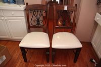 Pair of wooden dining chairs side by side showing metal scrollwork backs and white seats.