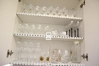 Three white shelves inside a cabinet holding assorted clear glass wine glasses and barware items.