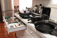 Various cookware, pots and pans including Wolfgang Puck pot, non-stick frypans, cast iron grill frypan, and kitchen drawer with utensils.