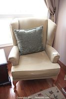 Front angle view of beige fabric wingback armchair with green cushion placed on wooden floor near window.