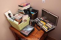 Overall view of multiple media types including baskets with CDs/DVDs and a recording case with cassette tapes on a wooden table.