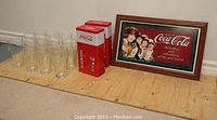 Set of 12 Coca-Cola glasses next to two Coca-Cola branded tins and a framed sign on the floor.