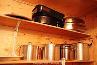Photo of two wooden shelves holding various covered pots and pans including several stainless steel pots and cast iron and enamel roasting pans.